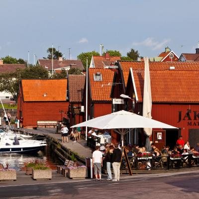 Röda små hus intill hamn i hamnstad. Restaurang med uteservering i förgrunden.