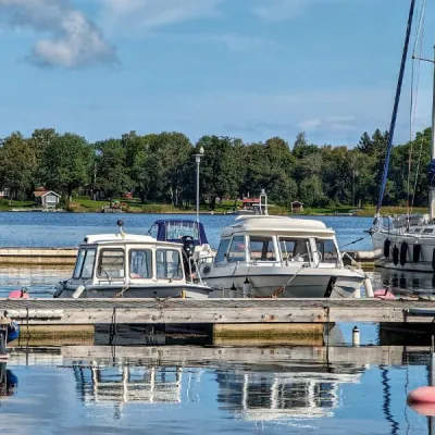 Båtar i hamn. Blått hav och grön skog och röd stuga på ö i bakgrunden