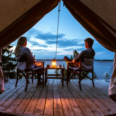 Öppningen i ett glampingtält mot terassen utanför där två personer sitter och titta på solnedgången över havet.