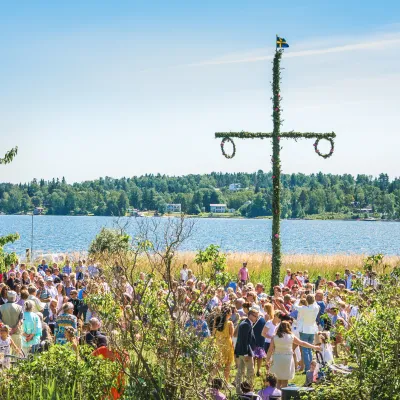 Midsommarfirande med en folkmassa runt en midsommarstång. Blå vattenspegel i bakgrunden och soligt väder.