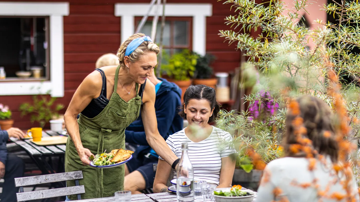 Uteservering framför röd stuga. Två gäster som blir serverade färgglad mat av servitris