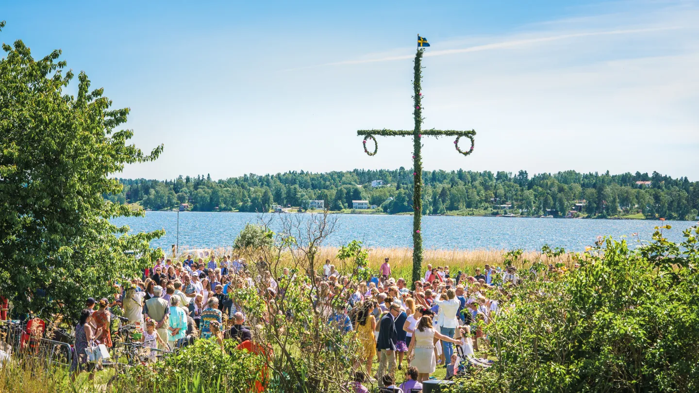 Midsommarfirande med folksamling kring midsommarstånd en solig dag med en blå vattenspegel i bakgrunden.