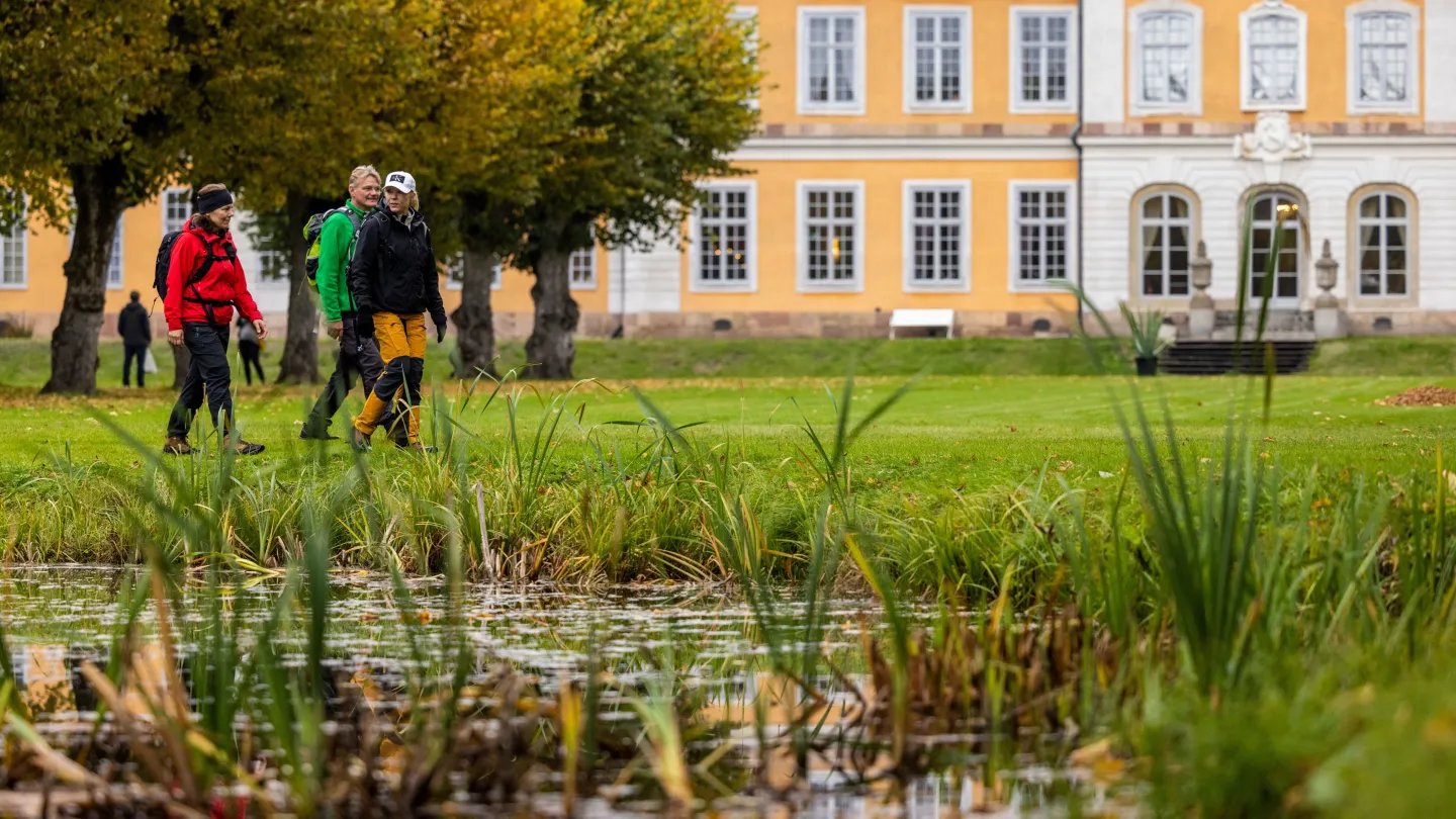 Tre höstklädda vandrare i en herrgårdsträdgård med herrgården i bakgrunden och en damm i förgrunden