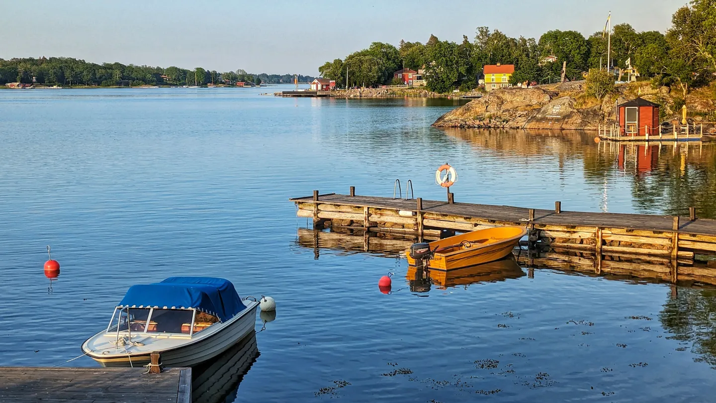 Båt vid brygga i förgrunden. Blått hav och blå himmel. Gråa klippor, gul stuga och grön skog i bakgrunden