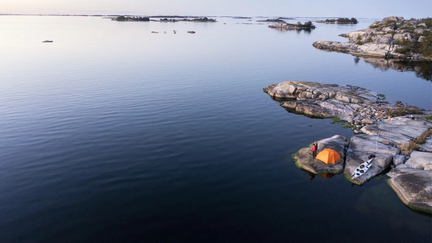 Drönarbild över ytterskärgården kring Norrpada. Blått hav, kajaker, kajakister och tält